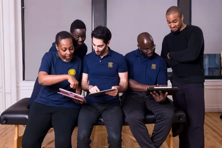 Group of students in matching navy blue uniforms collaborating on tablets and discussing notes, with a mentor observing and smiling in the background.